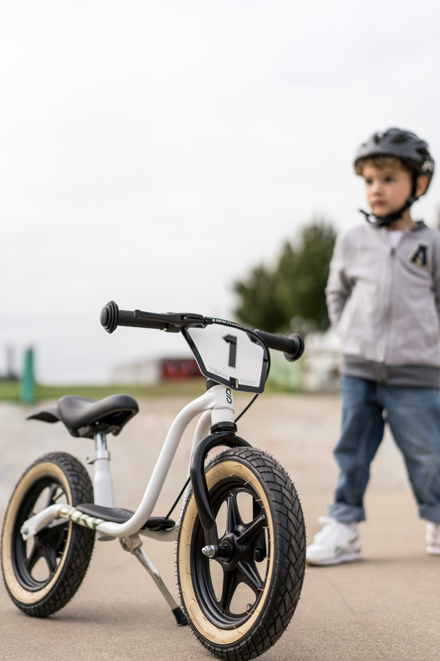 Ett litet barn med hjälm och grå jacka står bredvid en PUKY Balanscykel - Lufthjul - Puky LR 1_L Supermoto. Den vita cykeln har stora svarta pneumatiska däck, styre och en liten platta nummer 1 på framsidan. Bakgrunden visar ett utomhusområde med en molnig himmel.