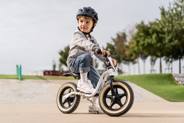 Ett litet barn med hjälm åker PUKY Balanscykel - Lufthjul - Puky LR 1_L Supermoto i en skatepark. Barnet är klädt i en grå luvtröja, jeans och vita sneakers. Träd och en betongramp syns i bakgrunden en molnig dag.