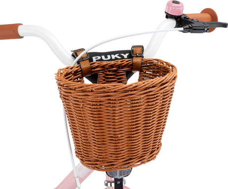 En närbild visar en PUKY Cykelkorg för 12" cyklar, en brun flätad korg, fäst vid styret på en balanscykel med vita handtag och rosa accenter, vilket ger charm och funktionalitet.